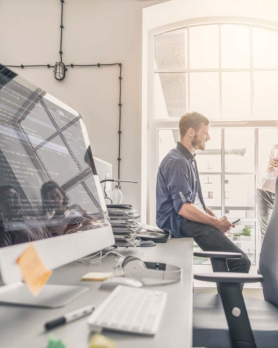 Helles Büro mit Bildschirmarbeitsplätzen und einer Besprechung im Hintergrund